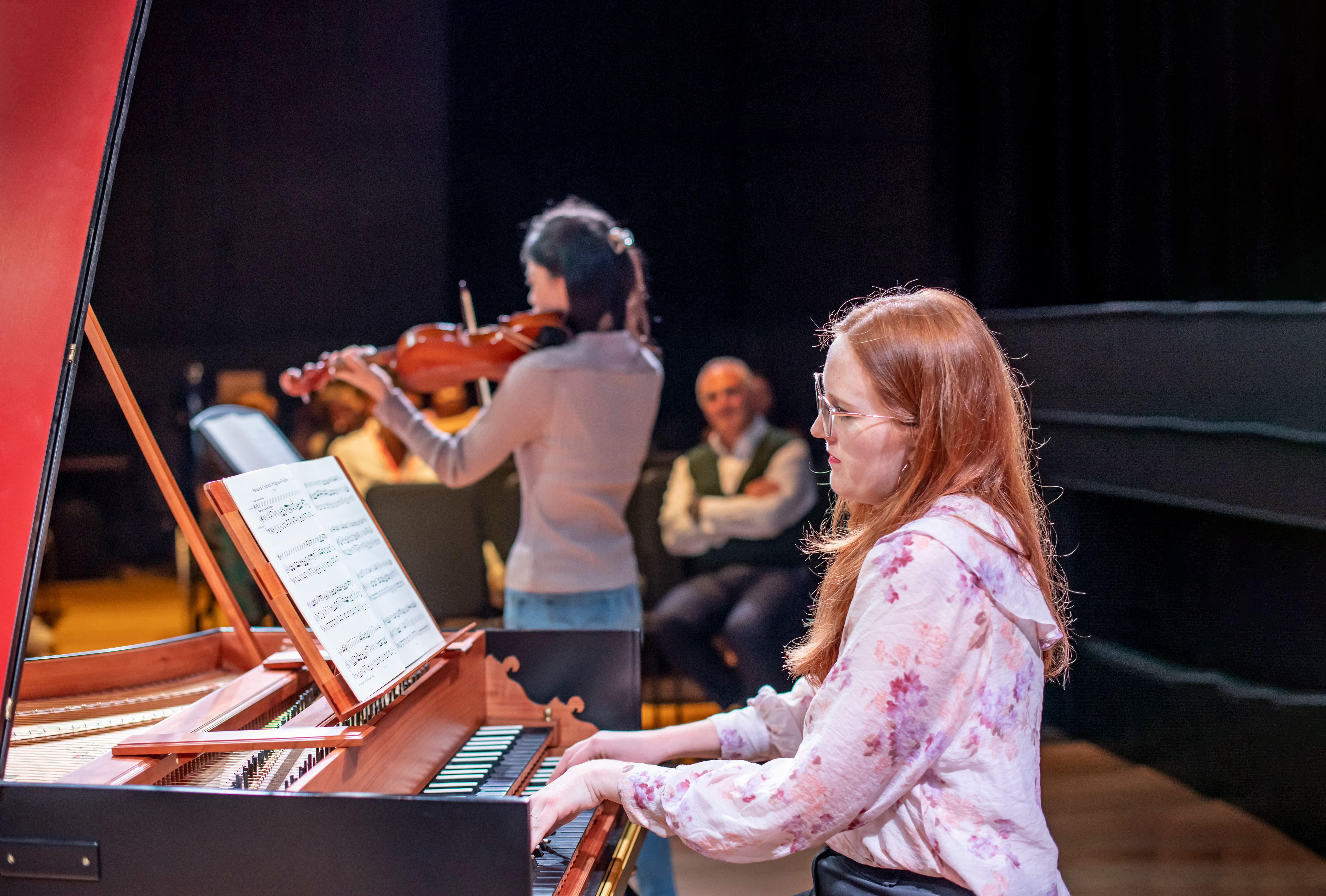 A female student, with glasses, red hair and a pink shirt, playing on a harpsichord, with a student performing in the background.